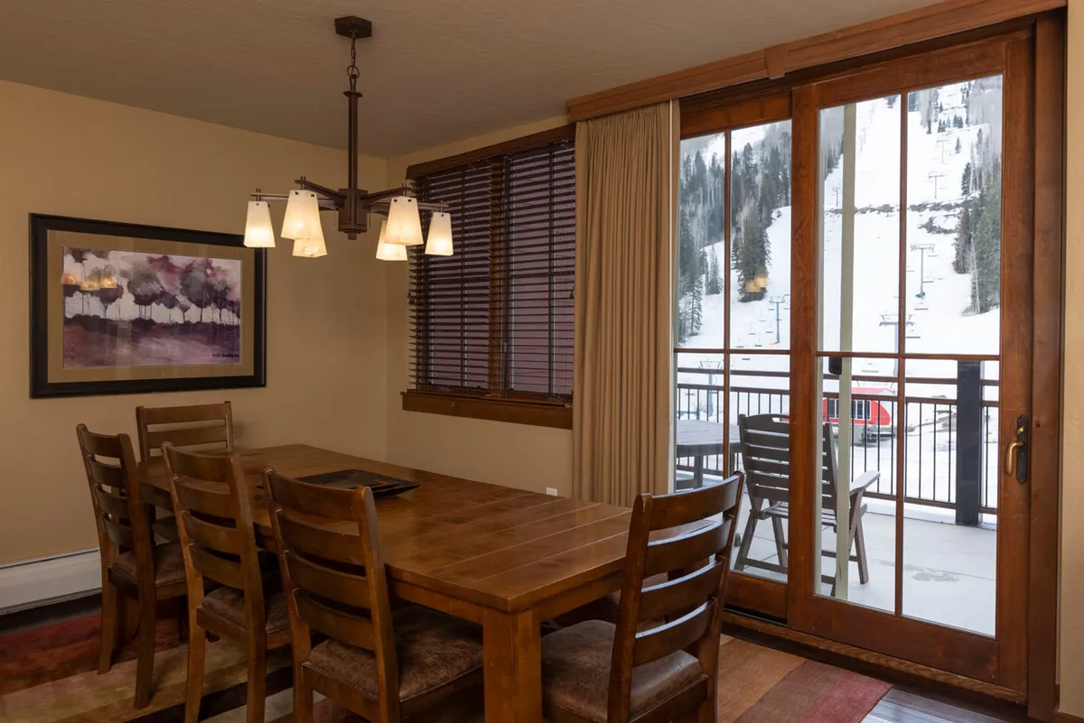 A bright dining area with a sturdy wooden table, elegant light fixture, and expansive windows offering views of a snowy mountain landscape. The large windows flood the space with natural light and create a seamless connection to the outdoors, enhancing the room's warm and welcoming atmosphere—ideal for family gatherings and scenic meals.