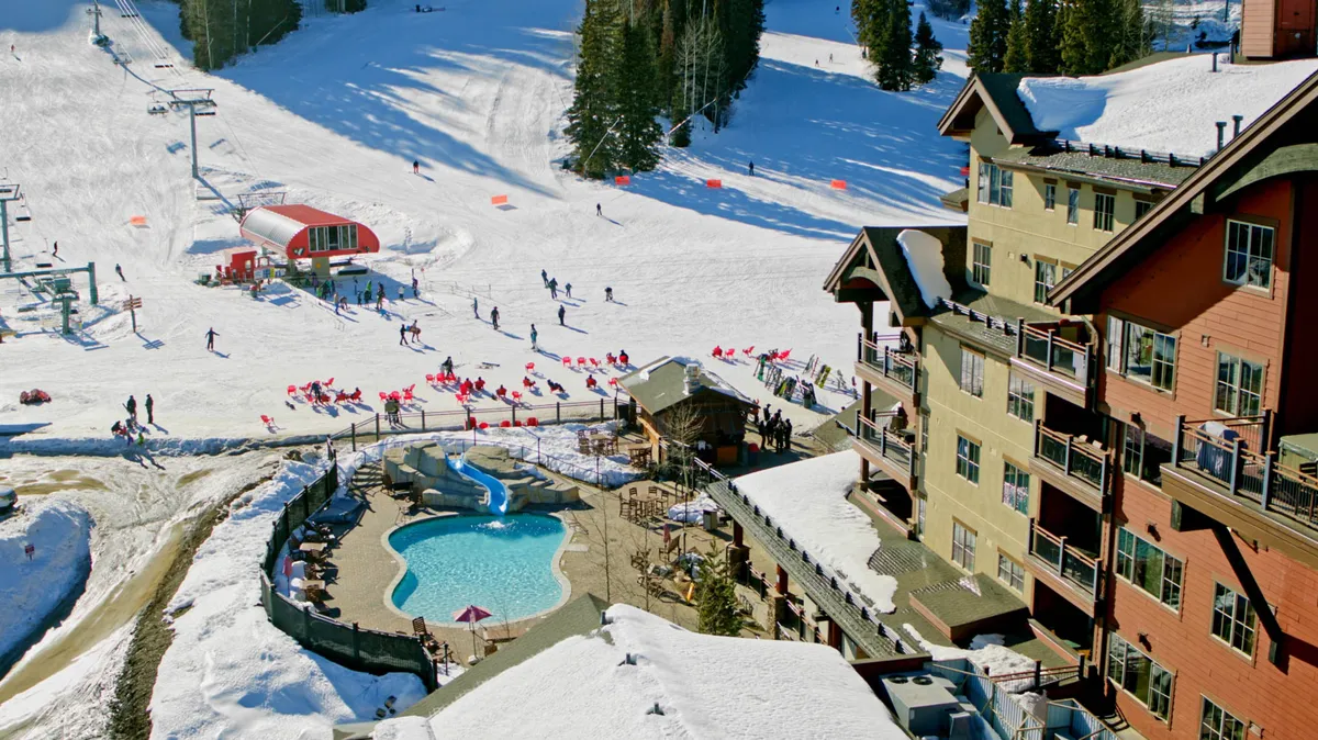 A vibrant exterior shot featuring the pool area with the ski slopes and lodge in the background. While the slopes suggest winter, the sunlit pool area evokes summertime relaxation, connecting directly to the mention of unwinding in the heated pool after a day of adventure.