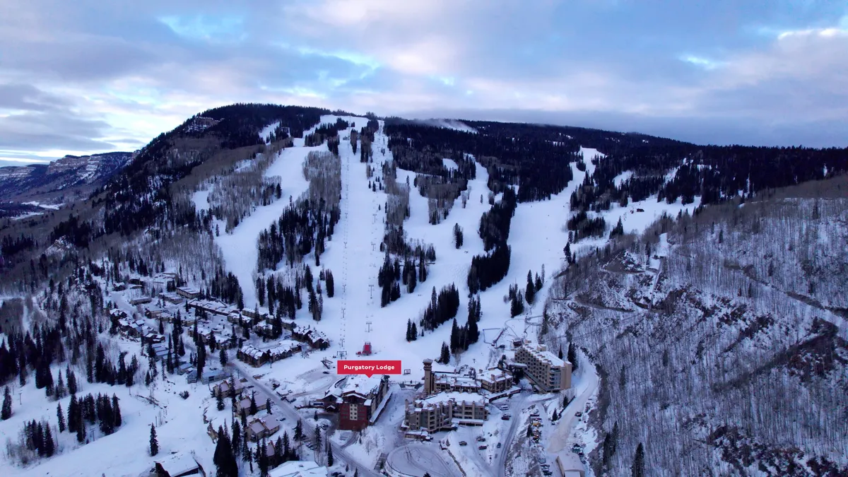 A panoramic view from near Purgatory Lodge, showing the snow-blanketed lodge, ski slopes winding down the mountain, and a forest of snow-tipped trees beneath a dramatic sunset sky. The warm lights of the lodge contrast with the cool snow, inviting guests in after a day outdoors.