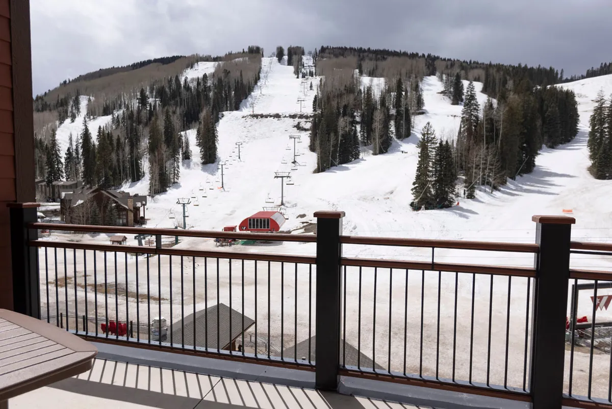 A wide-angle exterior shot capturing snowy ski slopes with fresh tracks, a ski lift in motion, and tall pine trees framed by a stunning mountain backdrop. The golden sunset bathes the landscape in warm hues, evoking the thrill of ski-in/ski-out access and the excitement of winter sports right outside your door.
