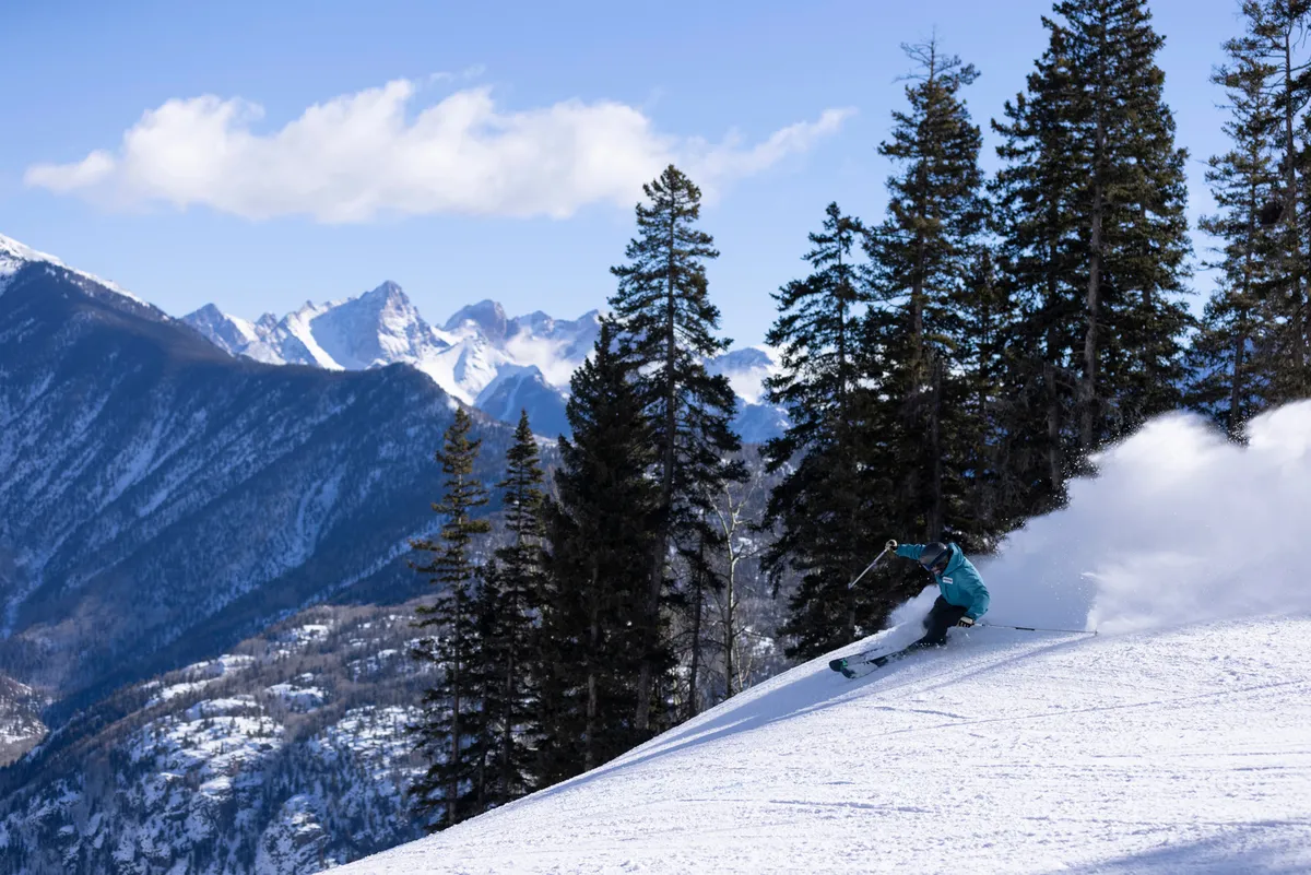A dynamic winter scene featuring a skier carving down a snow-blanketed slope, surrounded by pine trees and framed by distant mountains. The energetic action of the skier suggests adventure and fun, while the expansive natural setting highlights the resort’s access to outdoor recreation.
