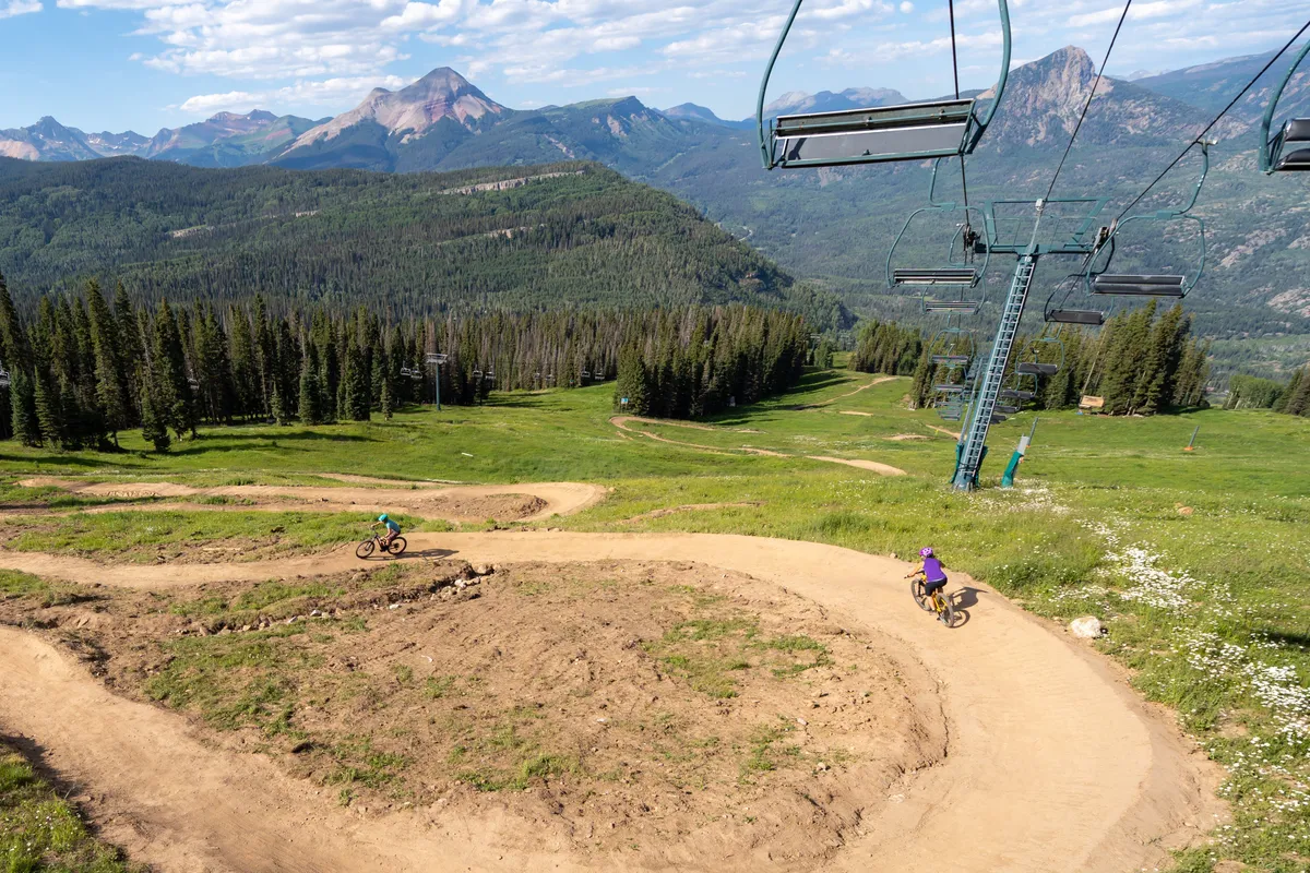 A sweeping exterior shot captures the mountain range under clear summer skies, with visible bike trails and a ski lift traversing the lush landscape. The scene evokes a sense of adventure, encouraging exploration by foot or bike and showcasing the natural beauty of the San Juan Mountains in the warmer months.