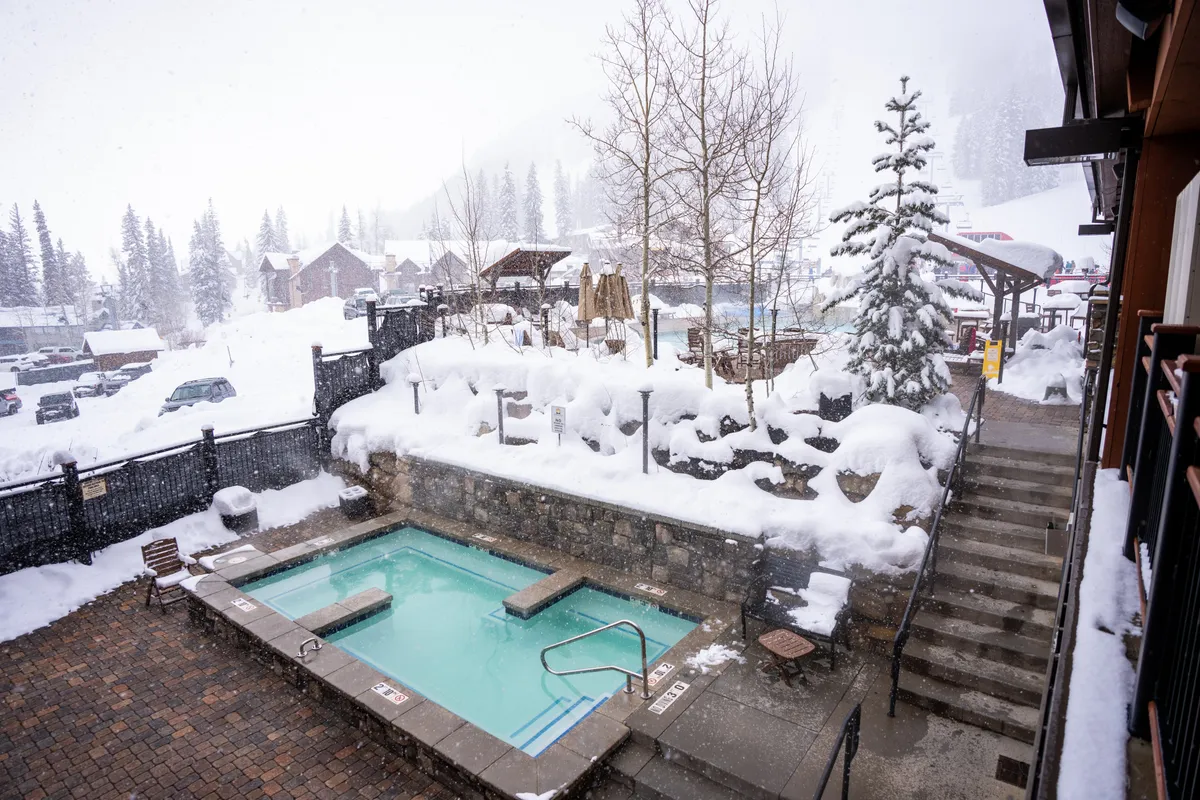 An inviting outdoor scene featuring a steaming hot tub nestled in a snow-covered landscape, surrounded by frosted trees. Steps lead up to the hot tub, suggesting easy access after a day spent on the slopes. The ambiance is tranquil, with soft evening light and hints of people relaxing and socializing.