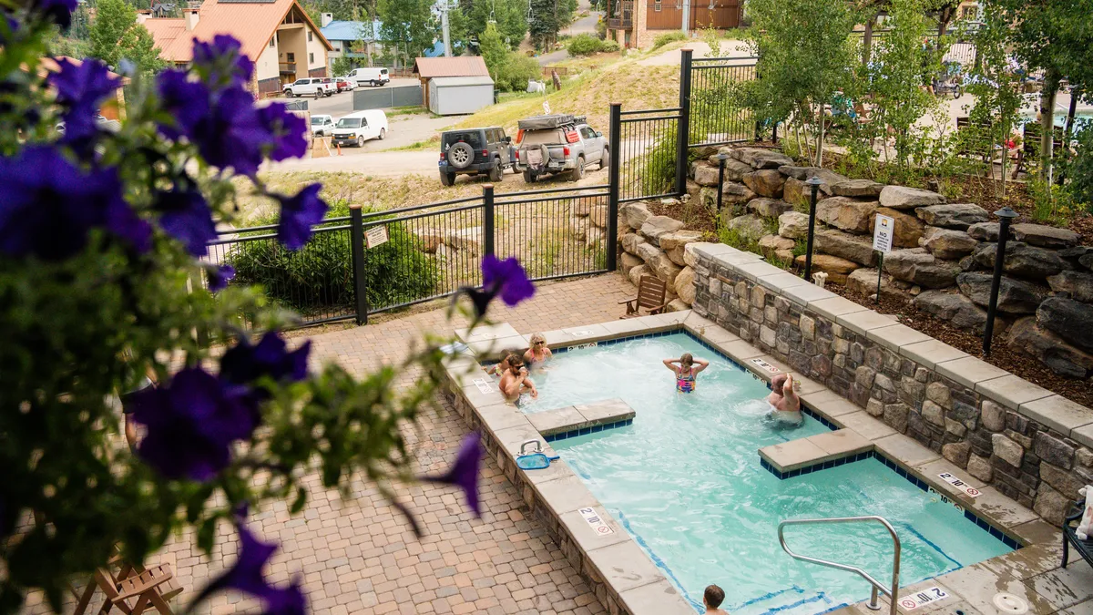 An inviting outdoor heated swimming pool with people enjoying themselves, surrounded by flowers and sun-soaked lounge spaces. The pool area provides a cheerful setting for relaxation, swimming, and family fun across all seasons.