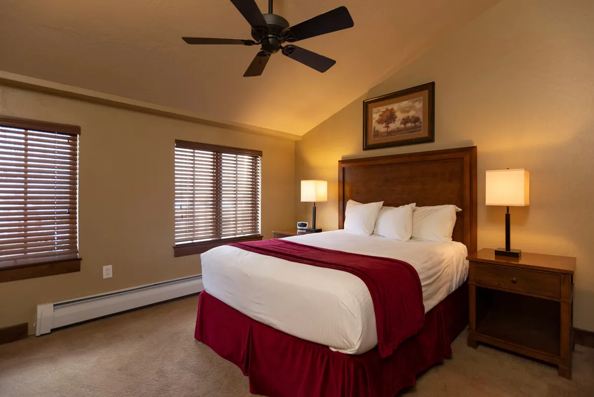A cozy interior bedroom scene featuring a plush bed with a red throw, wooden nightstand, wall art, and window blinds allowing gentle natural light to filter in. The room feels warm, inviting, and perfect for unwinding after a day of adventure or socializing.