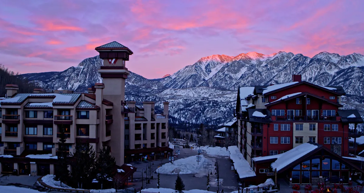 A sweeping exterior view of the ski resort buildings nestled at the base of a dramatic mountain range, bathed in the warm glow of a sunset. The snow-covered peaks and open sky evoke both adventure and tranquility, while the resort buildings suggest welcoming social spaces and amenities.