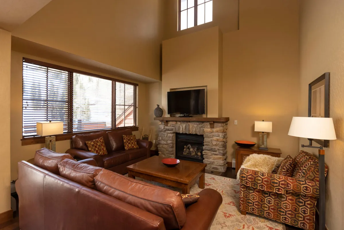 The image showcases a warm, inviting living space centered around plush leather sofas and a striking stone fireplace. Expansive windows frame breathtaking mountain views, flooding the room with natural light. The setup encourages gatherings, relaxation, and enjoying the comforts of a luxury condo in the heart of the resort.
