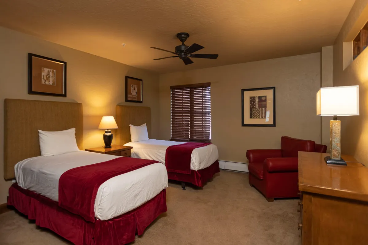 A cheerful bedroom with two twin beds, each featuring red accents, a bedside lamp, and a cozy armchair. The room is bright and inviting, ideal for children or extra guests. The setup emphasizes relaxation and a comfortable night’s sleep.